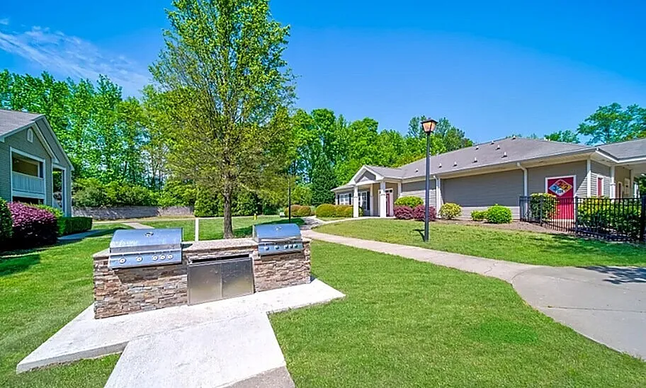 Charming outdoor community area with a modern grill station, lush green lawn, and inviting residential buildings under a clear blue sky.