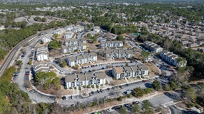 Aerial view of a picturesque apartment community surrounded by lush greenery and winding roads, showcasing modern residential buildings and ample parking.