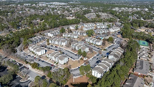 Aerial view of a modern apartment complex surrounded by lush greenery, showcasing a community with well-maintained buildings and ample parking space.