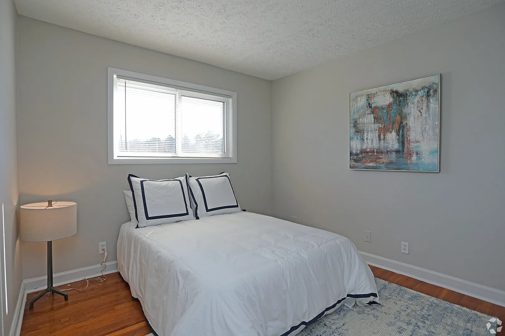 Cozy bedroom featuring elegant bedding, a modern table lamp, and stylish artwork, enhanced by natural light from a large window.