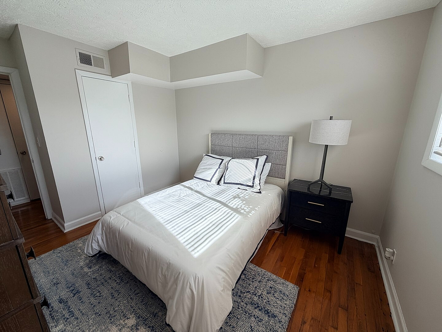 Bright bedroom with modern design featuring a cozy bed, elegant side table, and large window brings in natural light on hardwood floors.