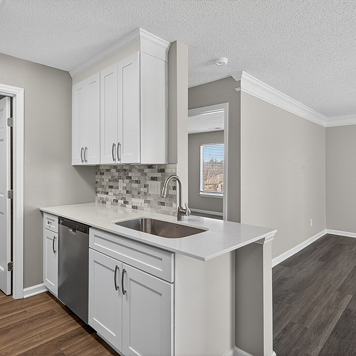 Modern kitchen with sleek white cabinetry, stylish tile backsplash, and elegant countertops, adjacent to a bright living area with wood flooring and large windows.