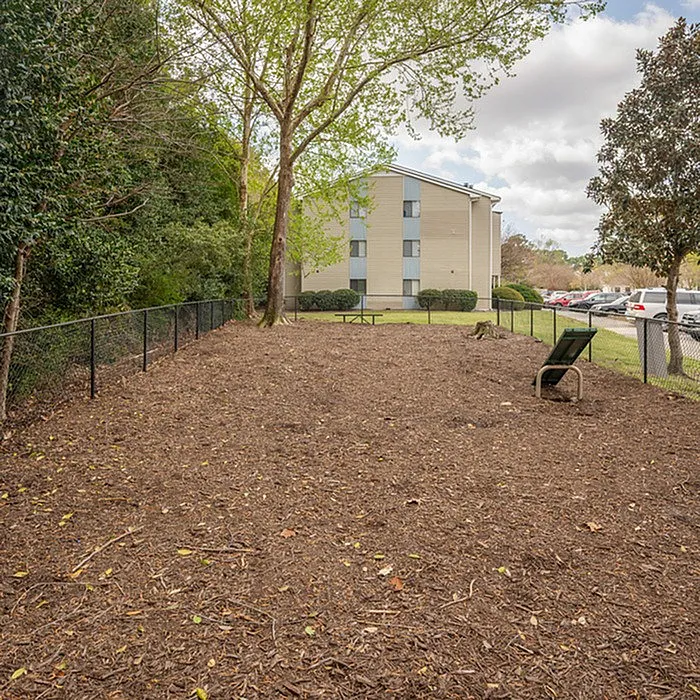 Inviting fenced-out dog park with bench seating and lush greenery surrounded by a charming apartment building.