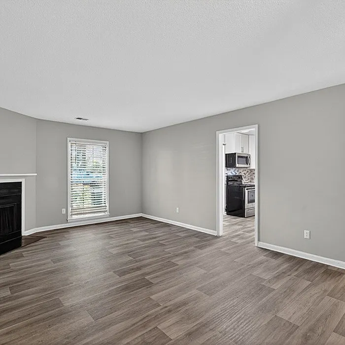 Inviting living room featuring modern hardwood flooring, ample natural light from a large window, and an open doorway leading to a contemporary kitchen.