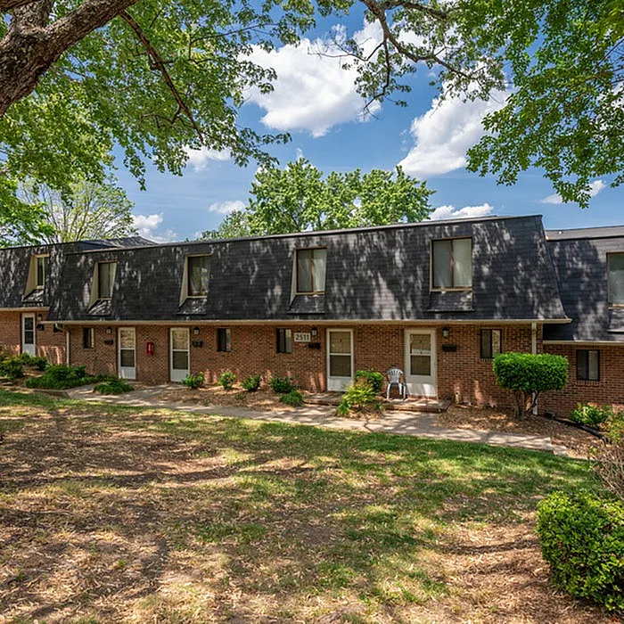 Charming brick townhouse nestled under lush, mature trees with inviting landscape and a serene atmosphere.