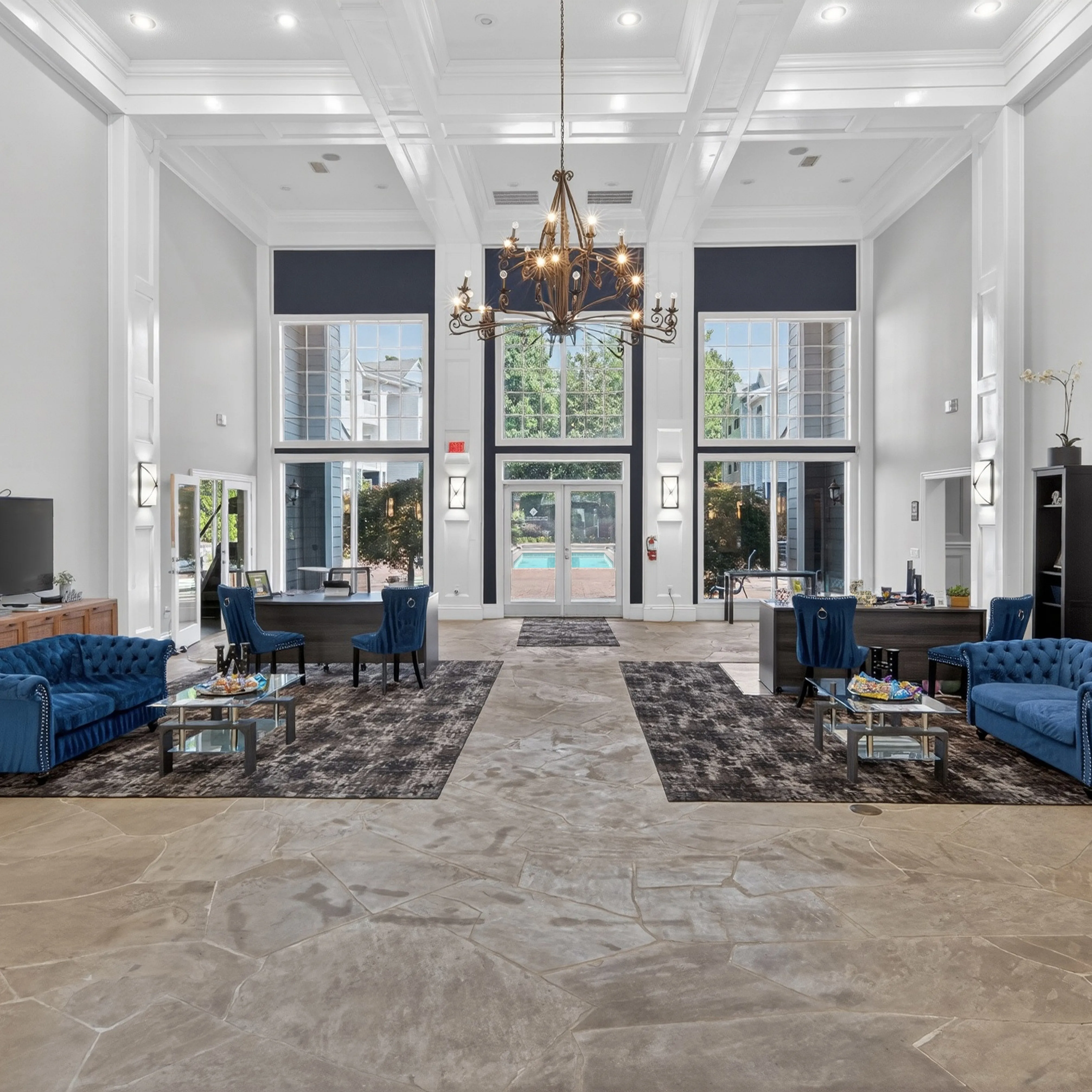 Luxurious apartment lobby with high ceilings, elegant chandeliers, plush blue sofas, and expansive windows offering a pool view.