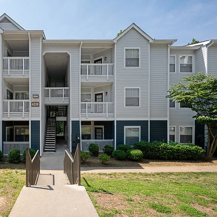 Charming apartment building with white siding and inviting balconies, surrounded by lush greenery and a clear path leading to the entrance.