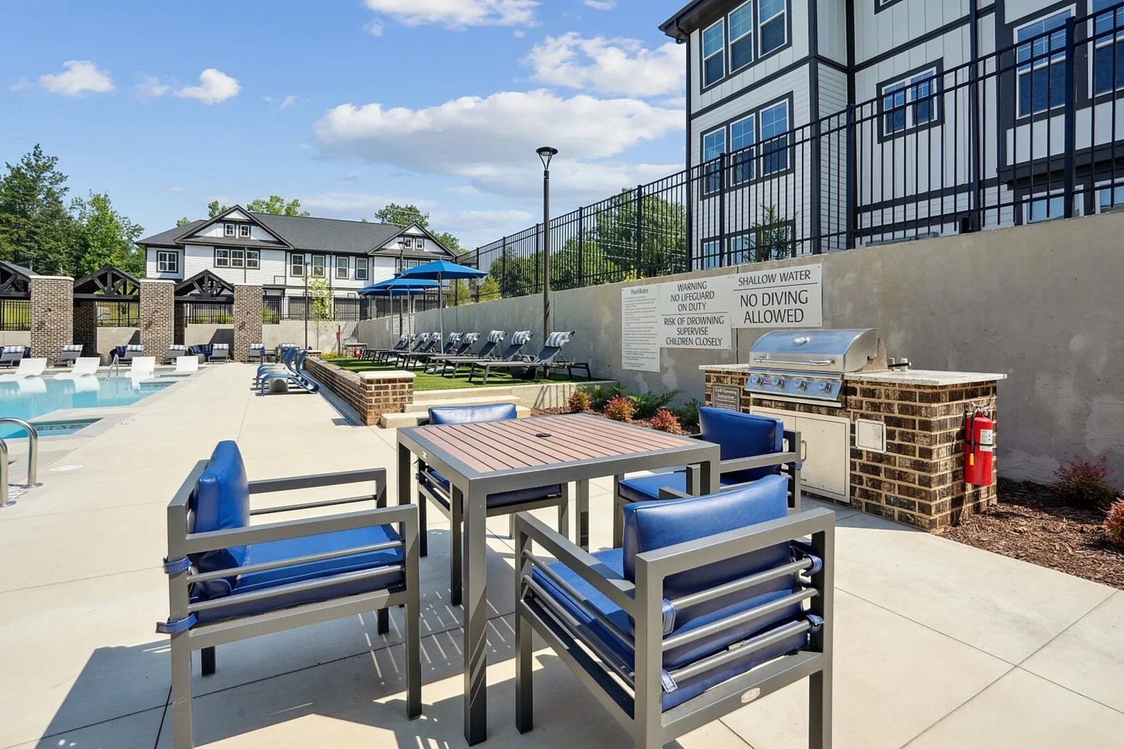 Luxurious apartment complex showcasing a sparkling outdoor pool, elegant patio seating, and modern barbecue area under a clear blue sky.
