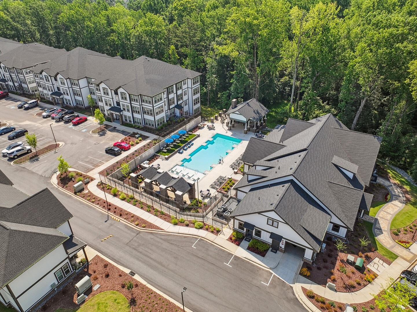 Aerial view of a modern apartment complex with a sparkling swimming pool, ample parking, and surrounded by lush greenery.