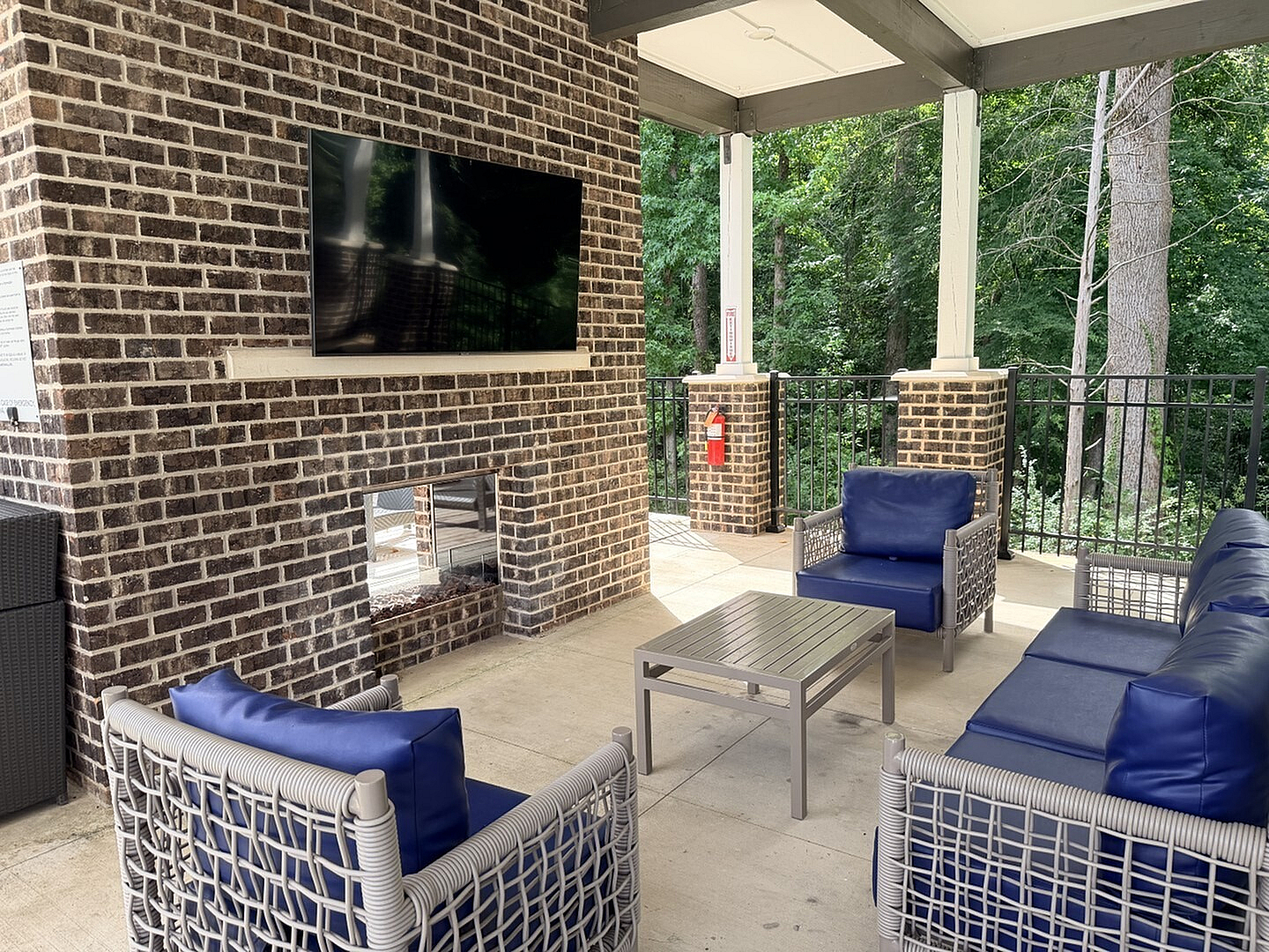 Chic outdoor lounge area with modern wicker furniture, blue cushions, a brick fireplace, and a flat-screen TV, surrounded by lush greenery.