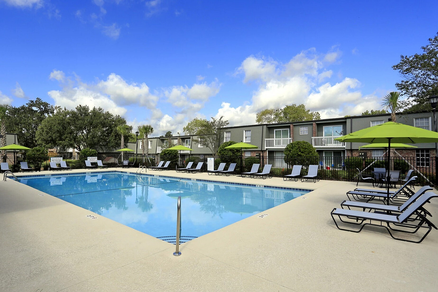 Inviting apartment complex with a large, sparkling swimming pool surrounded by comfortable lounge chairs and vibrant green umbrellas, perfect for relaxation and enjoying sunny days.
