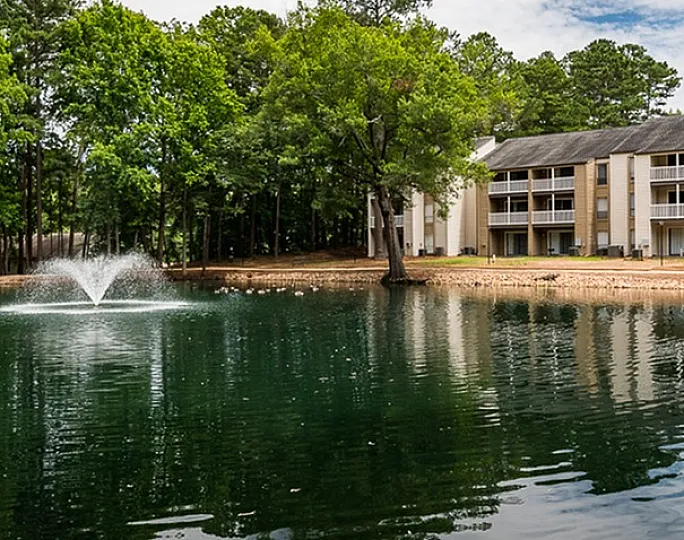 Charming apartment complex nestled by a tranquil lake with a serene fountain on a lush green backdrop.