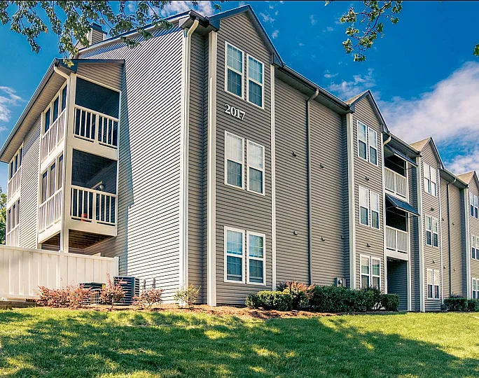 Modern three-story apartment building with balconies and manicured landscaping under a bright blue sky.