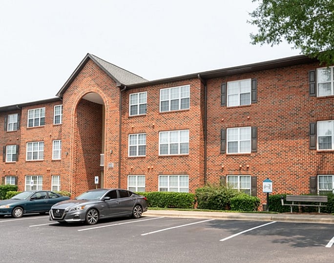 Red brick apartment building with spacious parking lot and lush greenery.