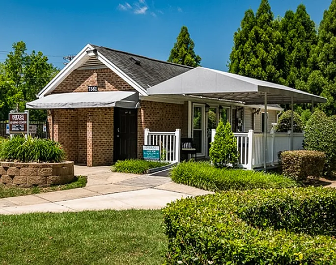 Quaint brick home with a welcoming porch surrounded by lush greenery and a clear blue sky.