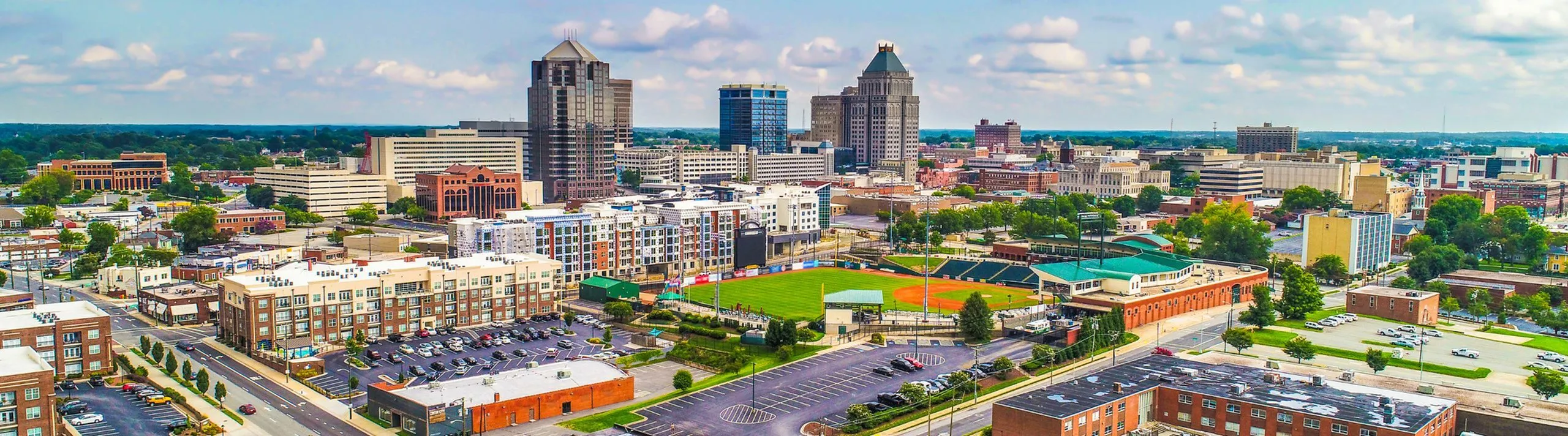 Panoramic aerial view of a bustling cityscape with modern high-rise buildings, spacious urban layout, and a prominent baseball field.