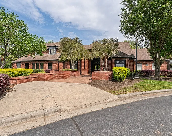 Charming brick apartment complex entrance with lush landscaping and mature trees, offering tranquility and elegance.