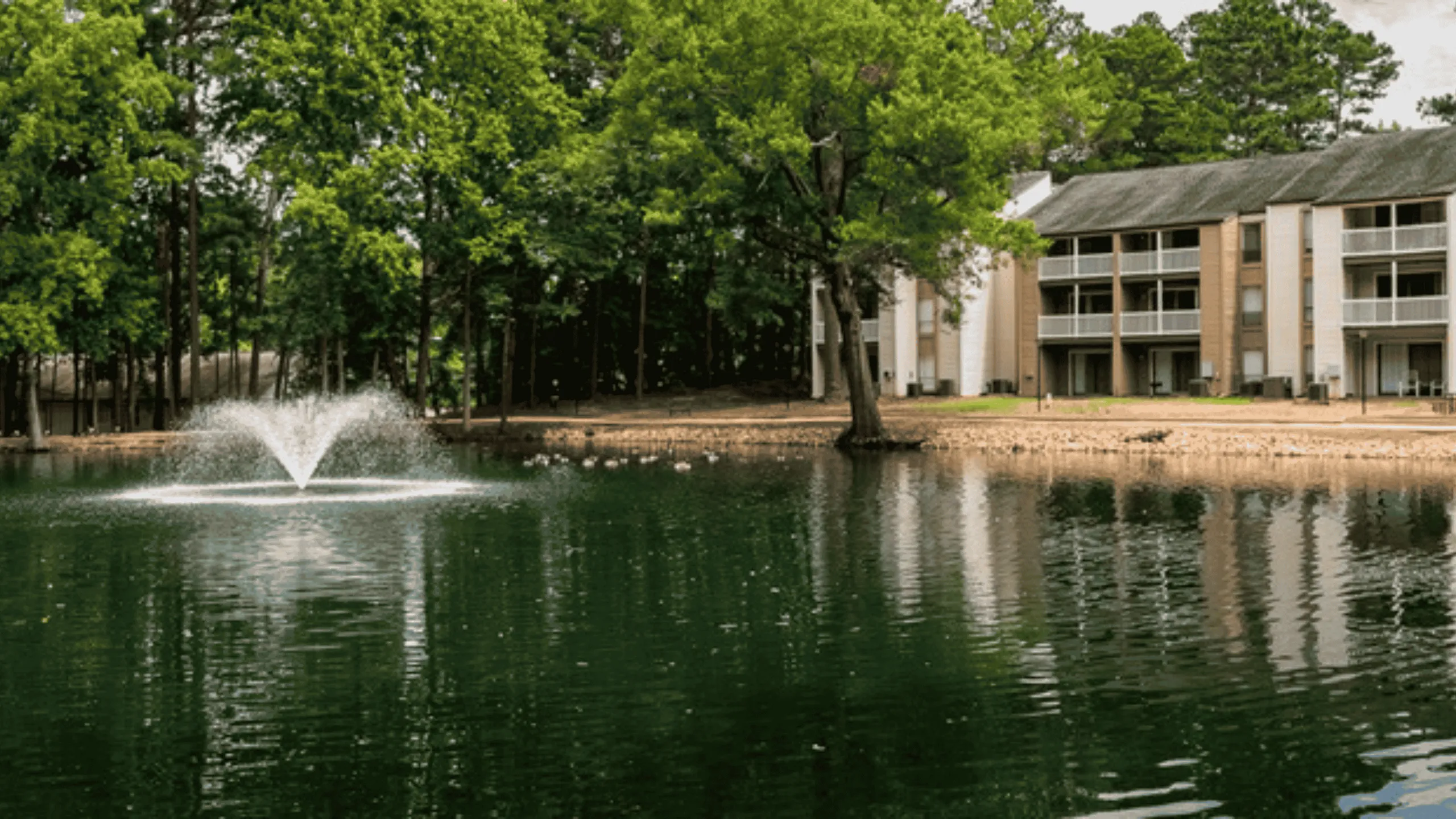 Serene apartment complex nestled by a picturesque lake with a charming fountain, surrounded by lush green trees.