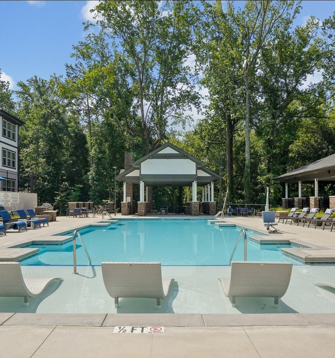 Luxurious outdoor swimming pool area with stylish lounge chairs, surrounded by lush greenery and modern apartment buildings.