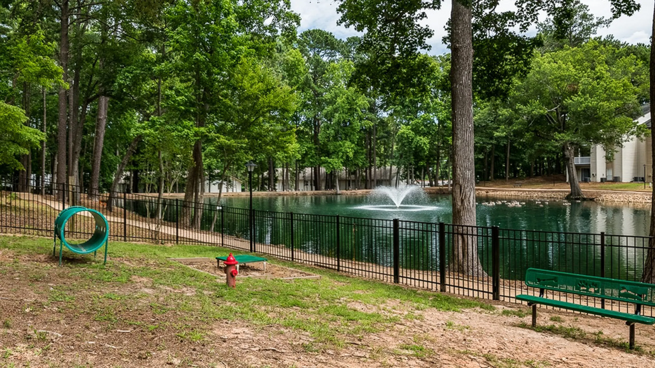 Tranquil outdoor area featuring lush trees, serene pond with fountain, and charming apartment view, perfect for relaxation.