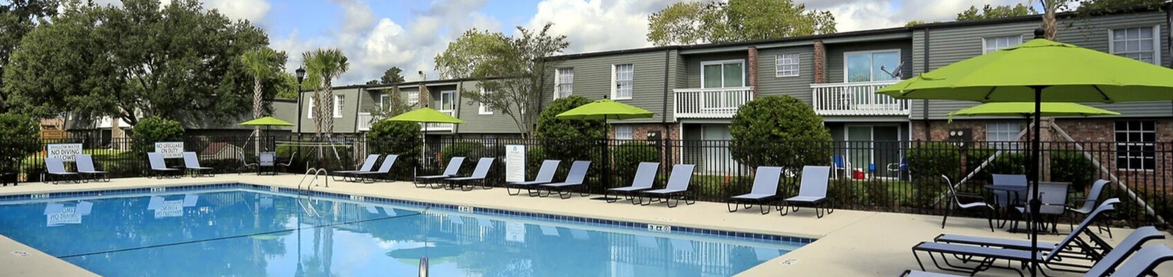 Inviting community pool area with lounge chairs and green umbrellas, surrounded by beautifully landscaped apartments under a bright blue sky.