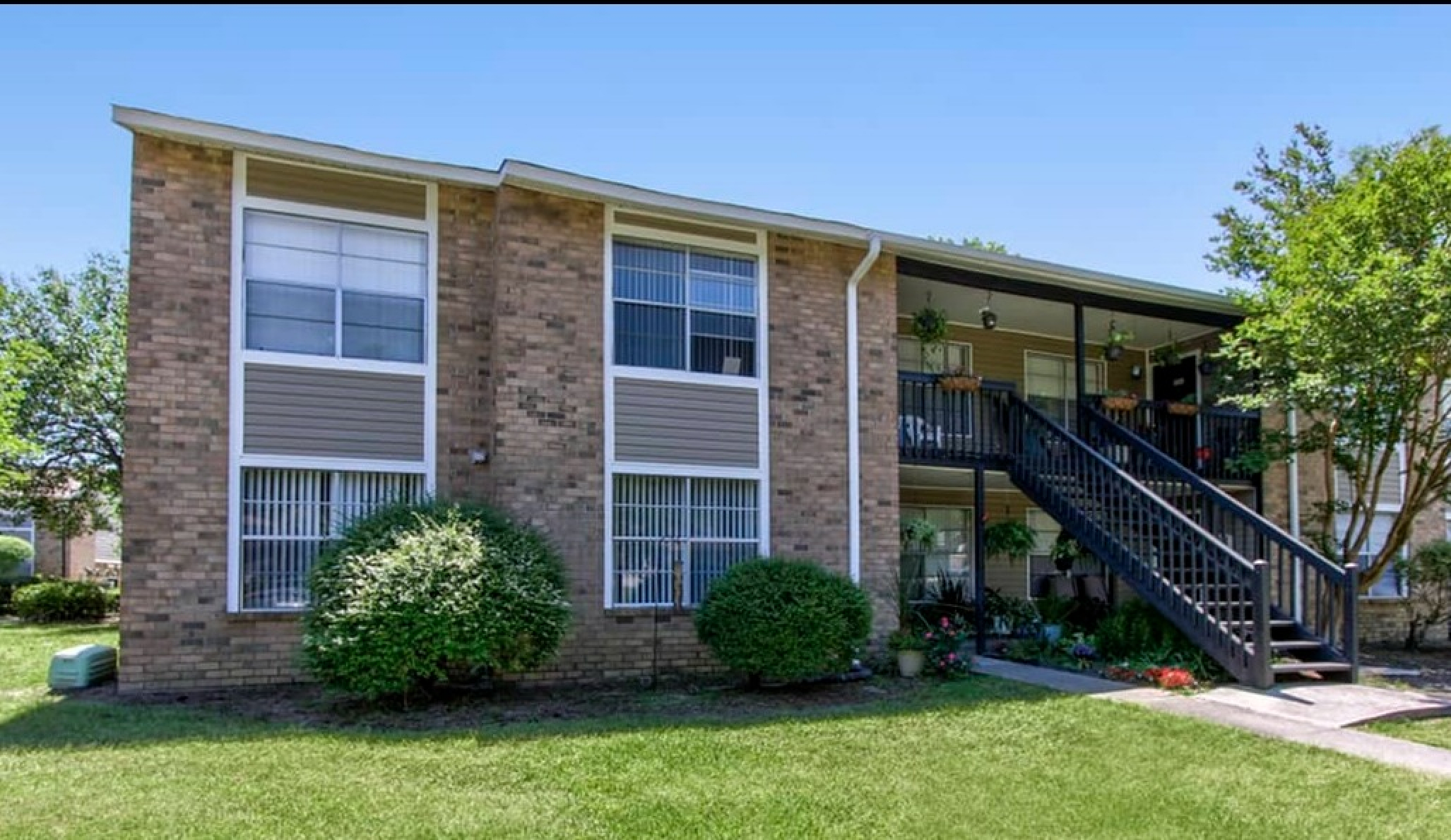 Charming brick apartment with lush greenery, inviting front garden, and spacious stairway leading to an open veranda.