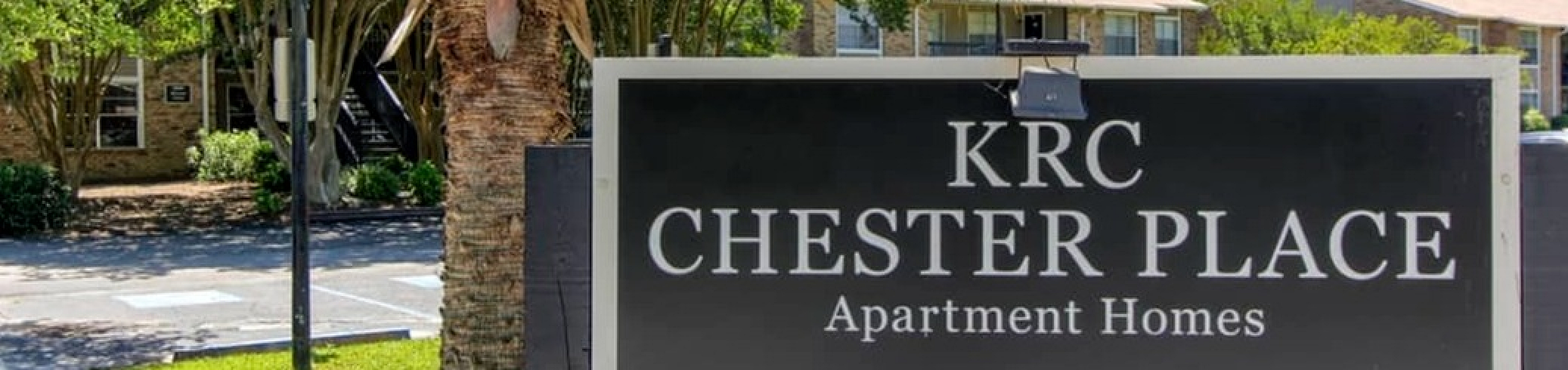Inviting entrance to KRC Chester Place Apartment Homes with lush palm trees and vibrant greenery.