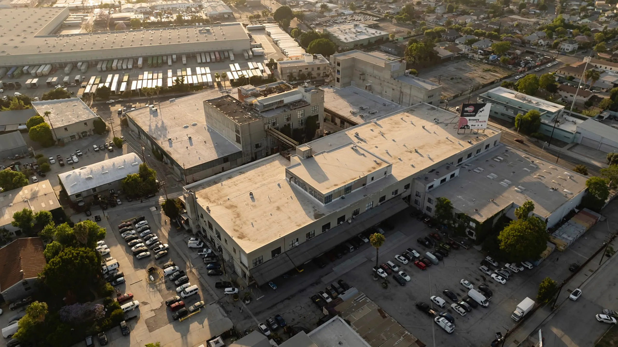 Aerial view of an expansive, sunlit industrial complex with ample parking and lush greenery, situated in a vibrant urban area.