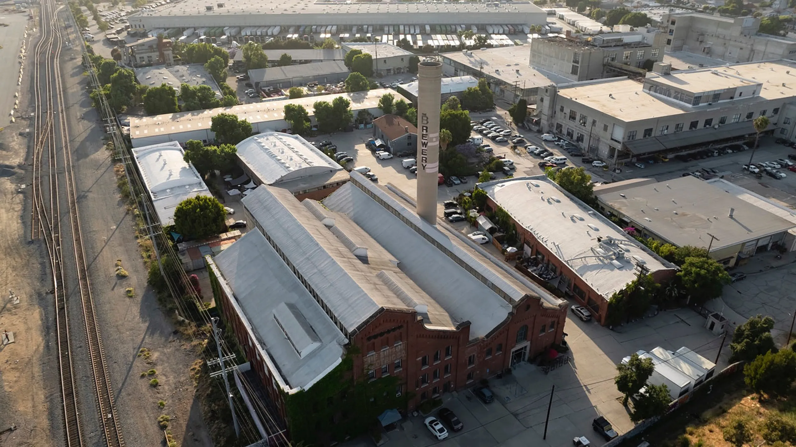 Aerial view of an expansive industrial complex featuring historic red brick architecture and greenery, adjacent to railway tracks, offering ample parking and vibrant surroundings.