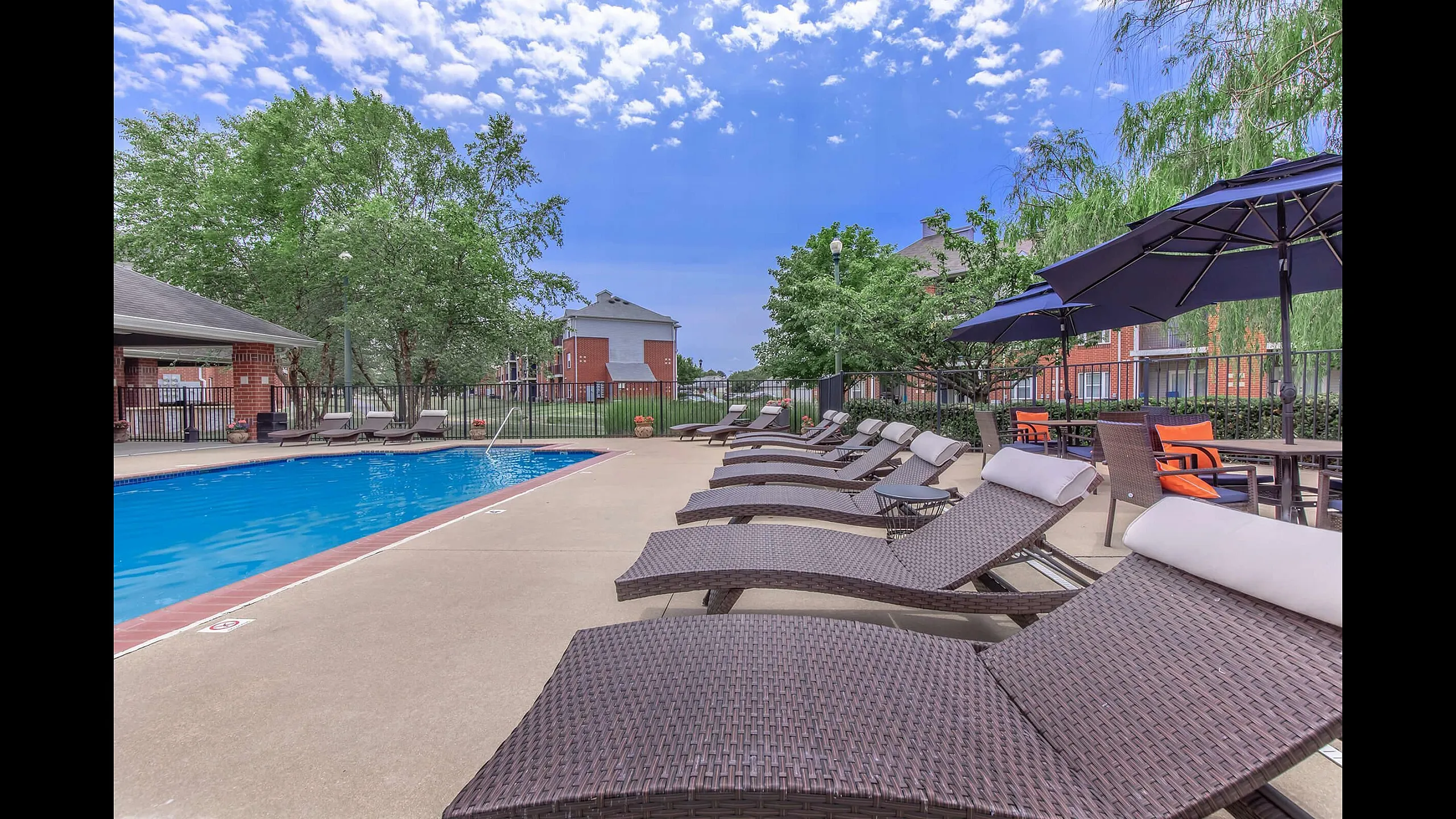 Luxurious outdoor pool area with stylish lounge chairs and vibrant umbrellas, surrounded by lush greenery and modern brick buildings under a clear blue sky.