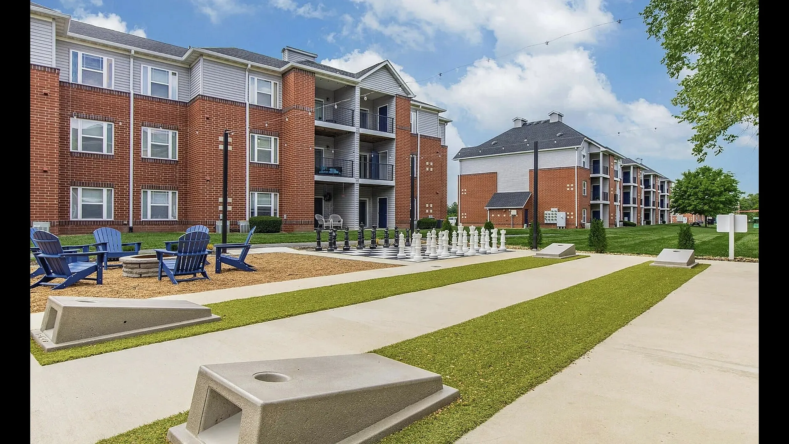 Inviting apartment complex with recreational amenities including a life-sized chess board, fire pit area, and cornhole games, set against a backdrop of charming brick buildings and lush green lawns.