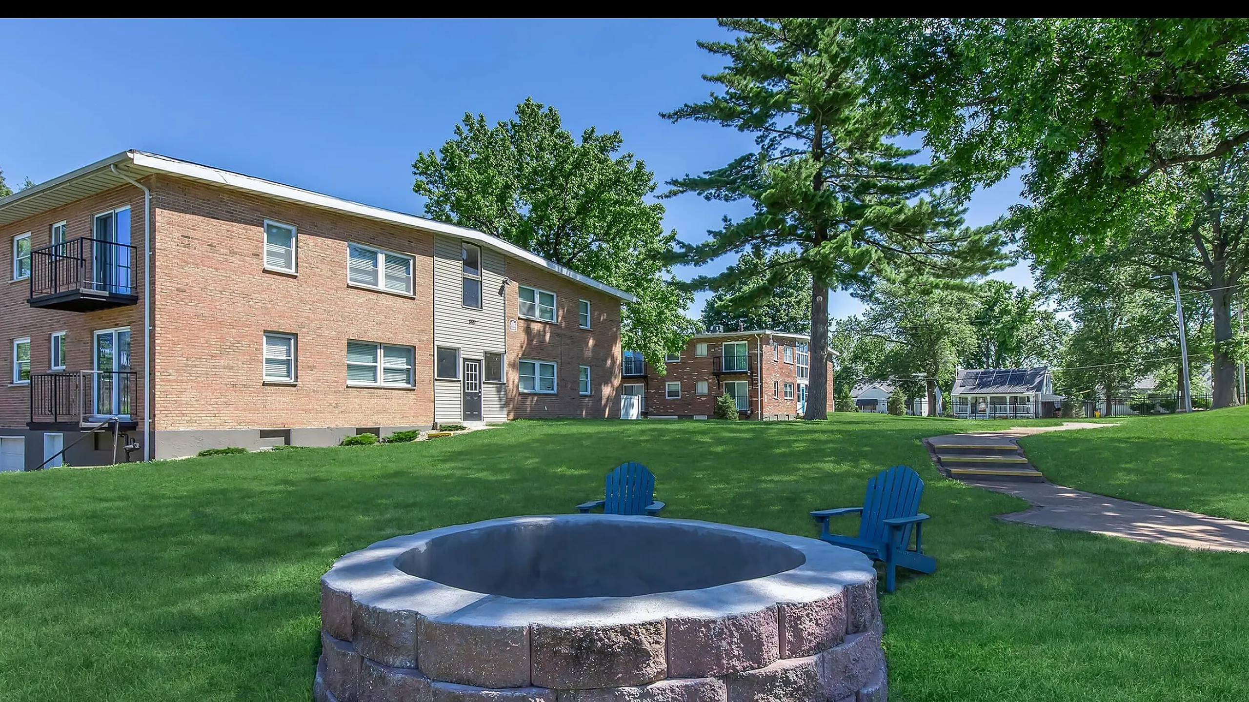 Charming brick apartment buildings surrounded by lush green lawns and mature trees, complete with a cozy fire pit and inviting blue Adirondack chairs.