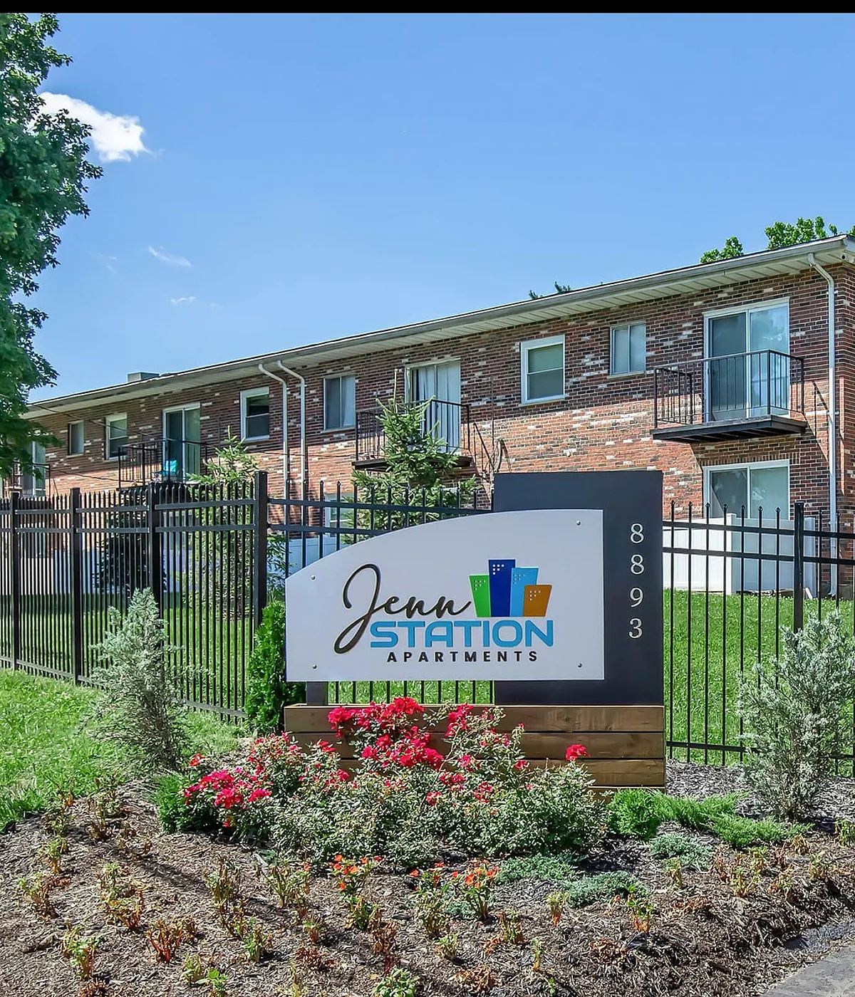 Charming Jenn Station Apartments showcasing a brick exterior with landscaped gardens and secure fencing under a bright blue sky.