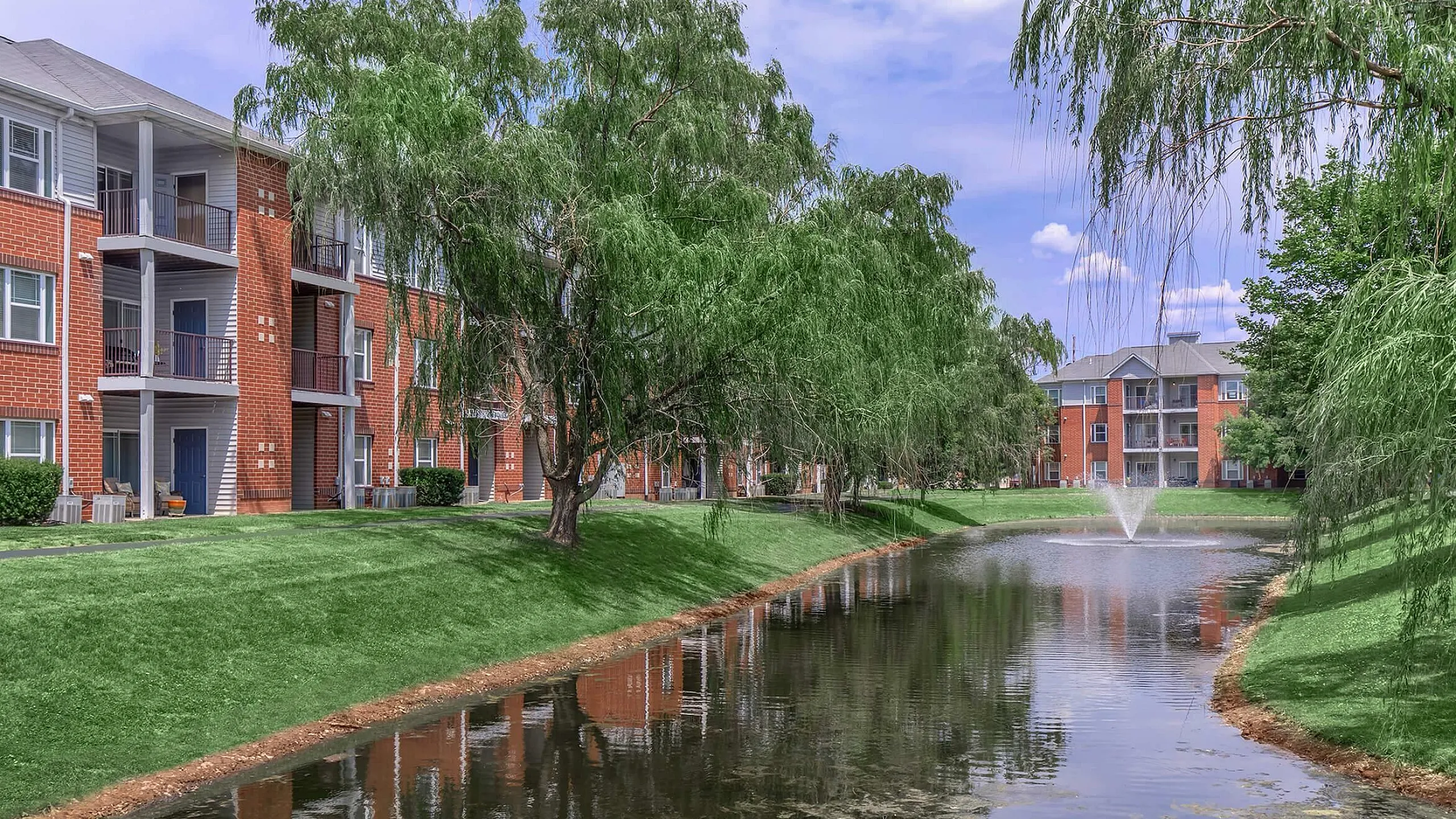 Charming apartment complex with brick exterior surrounded by lush greenery and a tranquil pond with a fountain.