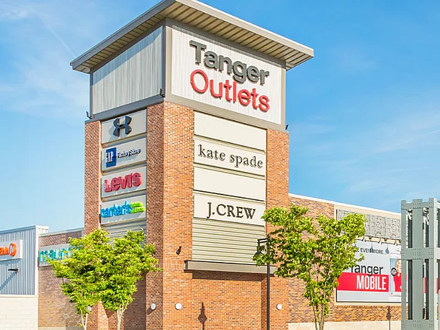Chic Tanger Outlets entrance showcasing popular brand signs, lush landscaping, and vibrant atmosphere under blue skies.