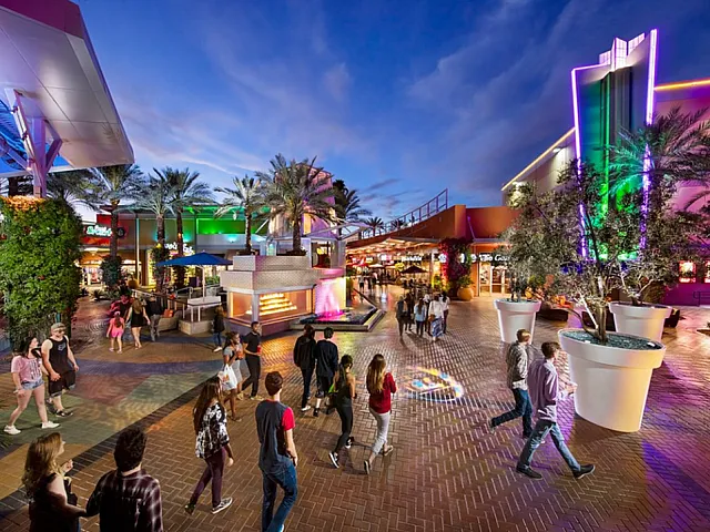 Vibrant night scene at an outdoor shopping and entertainment district with colorful lights, palm trees, and people strolling along the paved plaza.