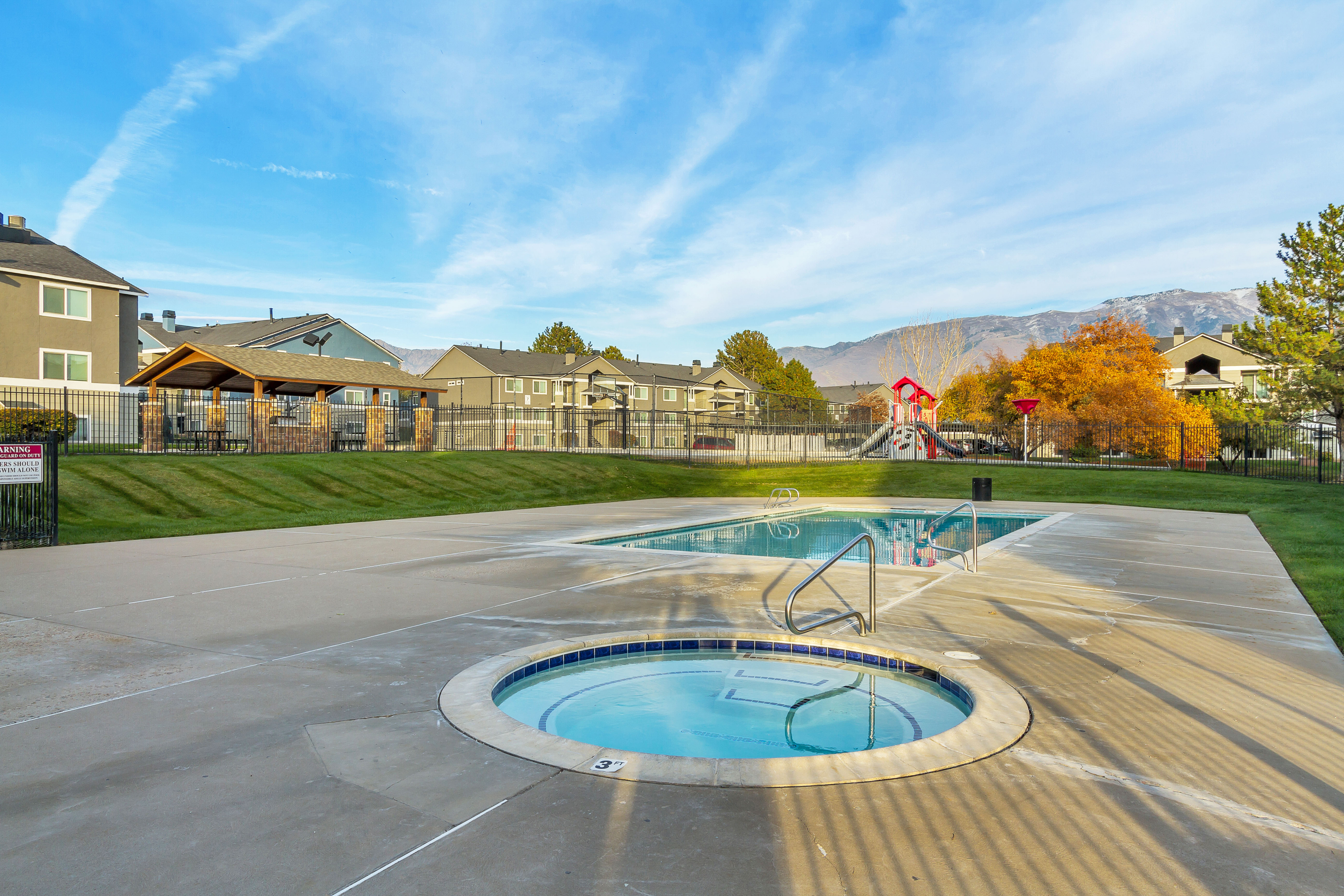 Inviting outdoor pool and hot tub area with scenic mountain views, surrounded by lush greenery and modern apartment buildings.