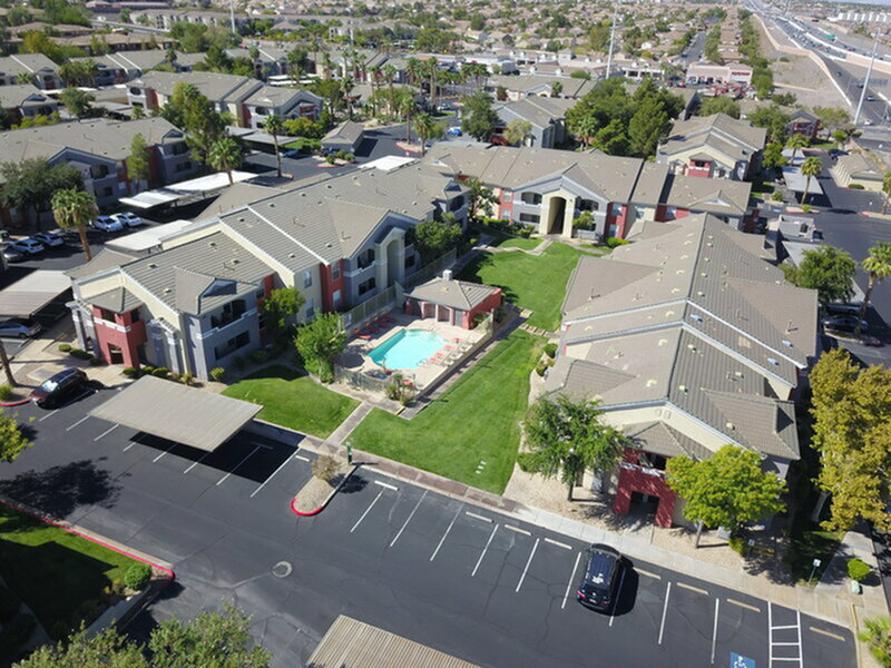 Aerial view of a modern apartment complex featuring a central pool, lush green landscaping, and ample parking spaces, nestled in a vibrant community.