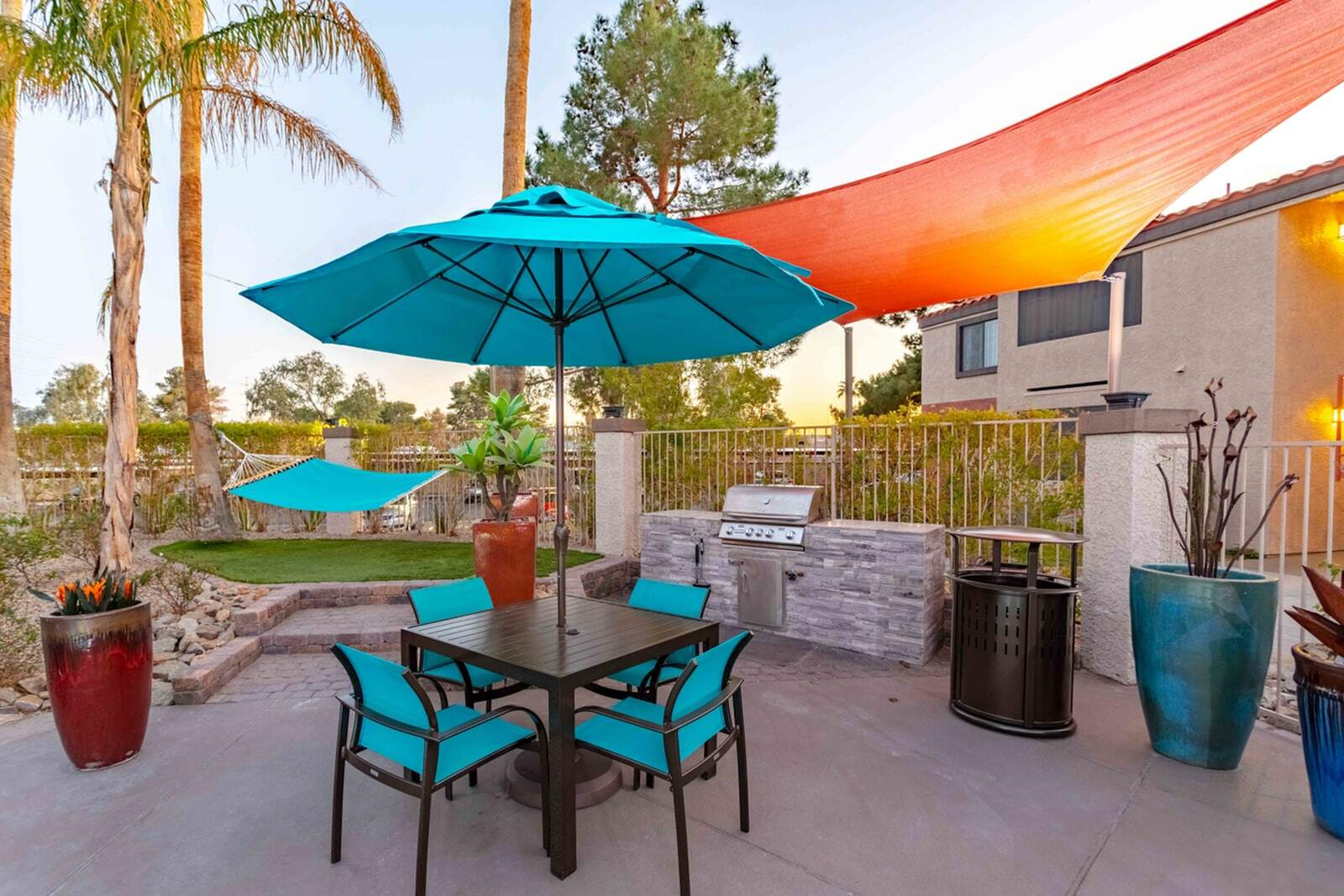 Vibrant patio with turquoise umbrella, sleek dining set, hammock, and modern BBQ grill, surrounded by lush greenery and palm trees.