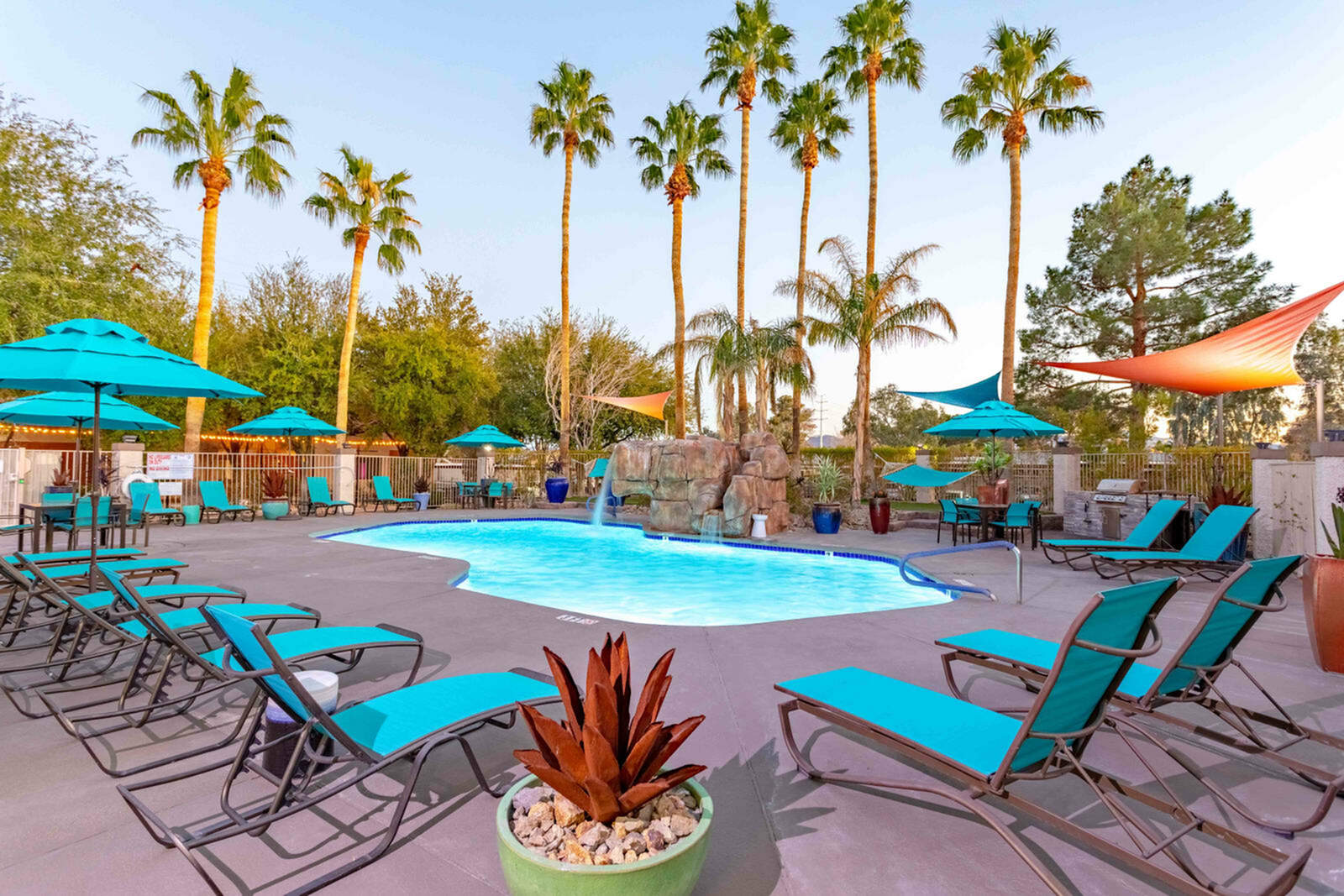 Luxurious outdoor pool area featuring vibrant turquoise sun loungers and umbrellas, surrounded by tall palm trees in a resort-style setting.