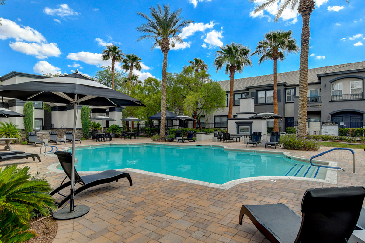 Luxurious apartment complex with a sparkling pool, elegant lounge chairs, and lush palm trees under a clear blue sky.