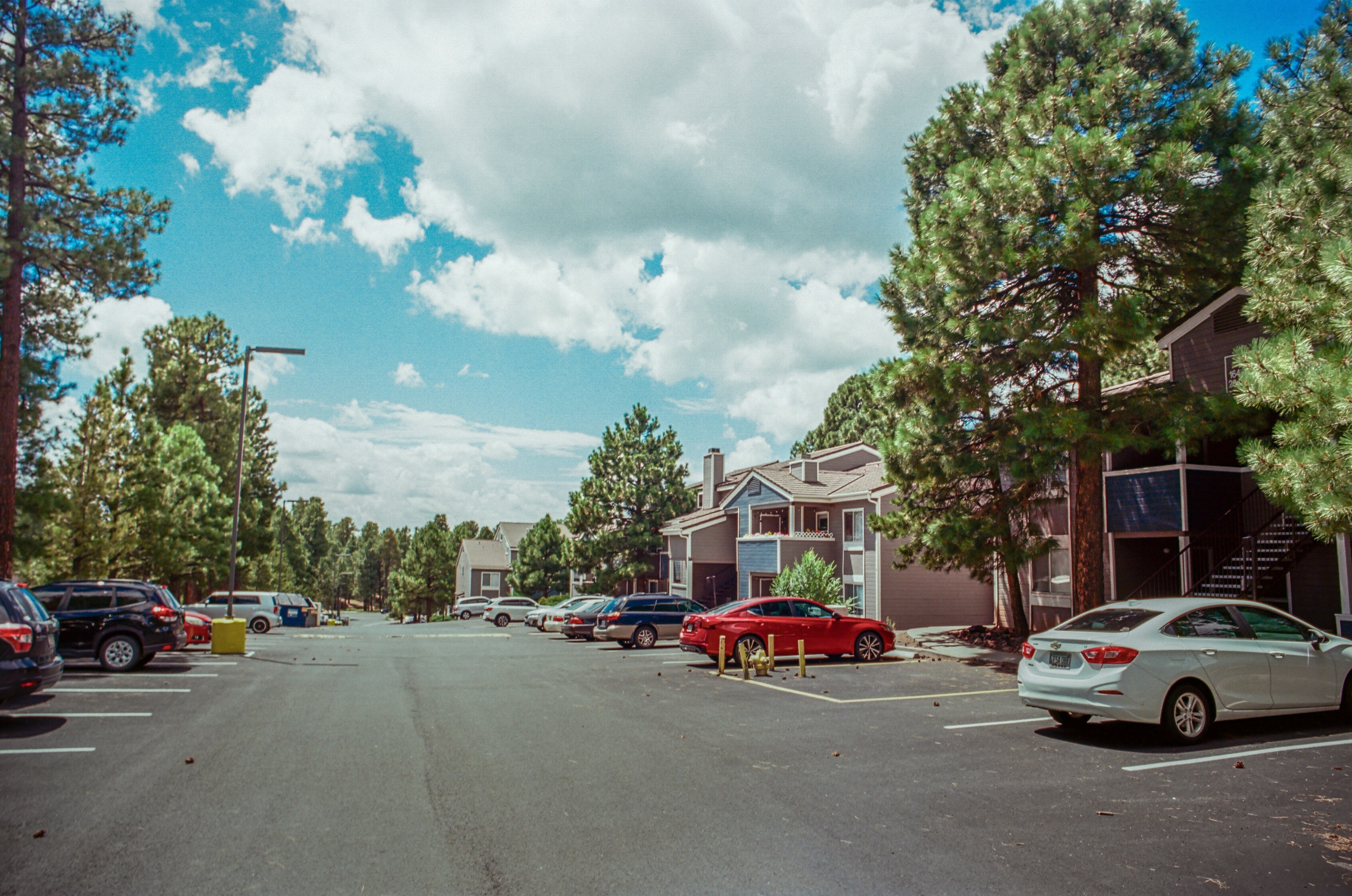 Charming apartment building surrounded by lush greenery and a clear blue sky, featuring ample parking space and serene outdoor environment.