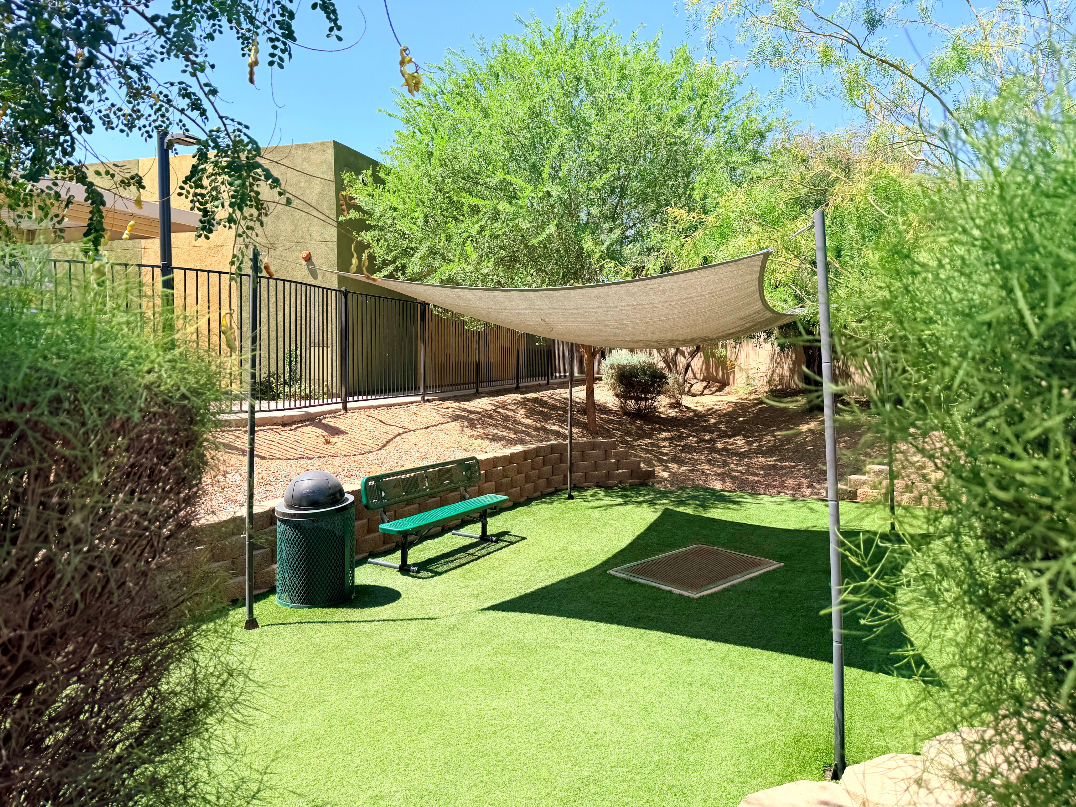 Charming outdoor area with lush greenery, a cozy bench under a sunshade, and modern fencing; perfect for relaxation.