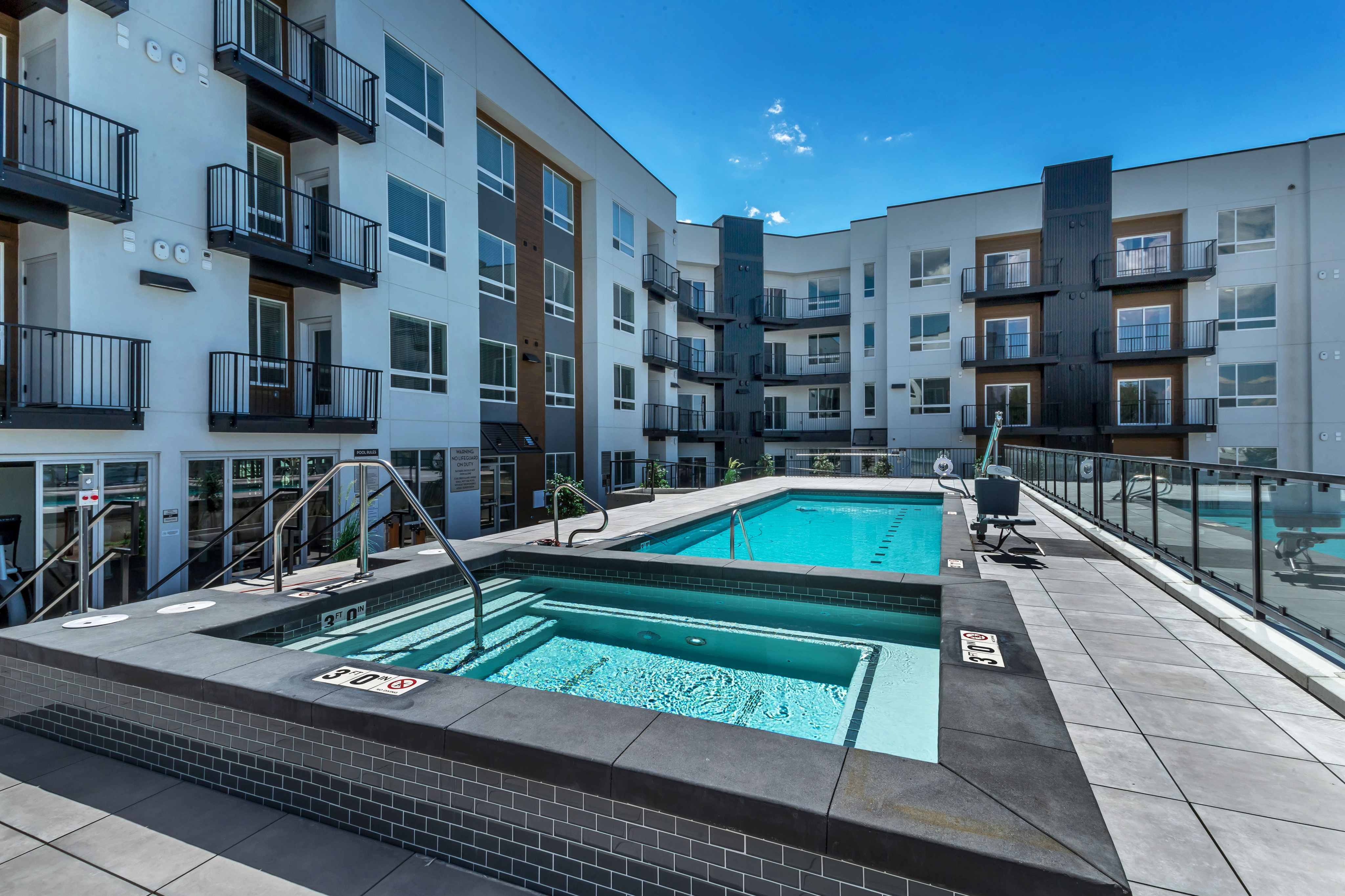 Modern apartment complex with a sparkling pool and hot tub, surrounded by stylish balconies and sleek architecture under a clear blue sky.