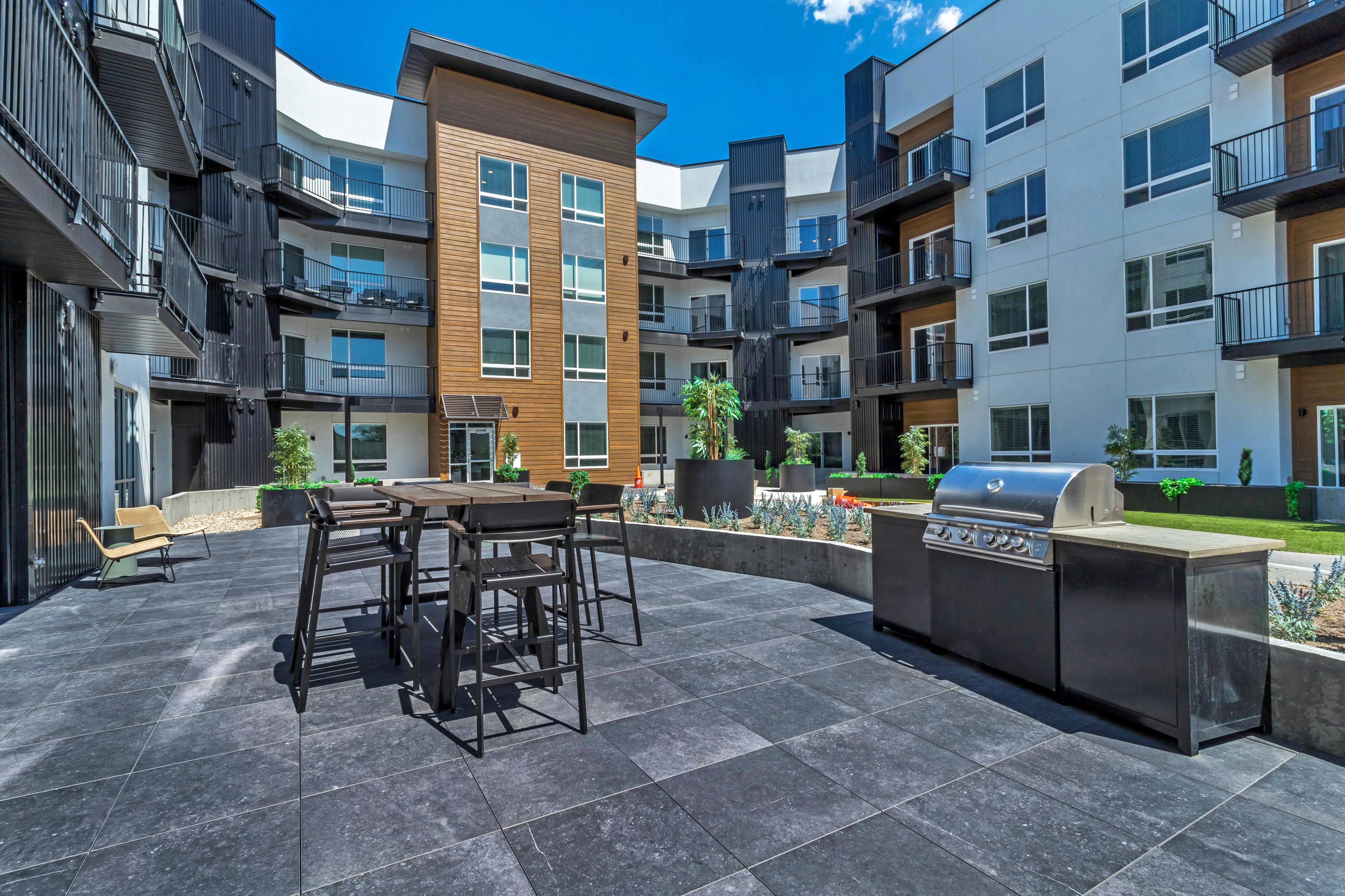 Modern apartment courtyard with sleek patio furniture, vibrant greenery, and a stainless steel grill set against stylish architecture.