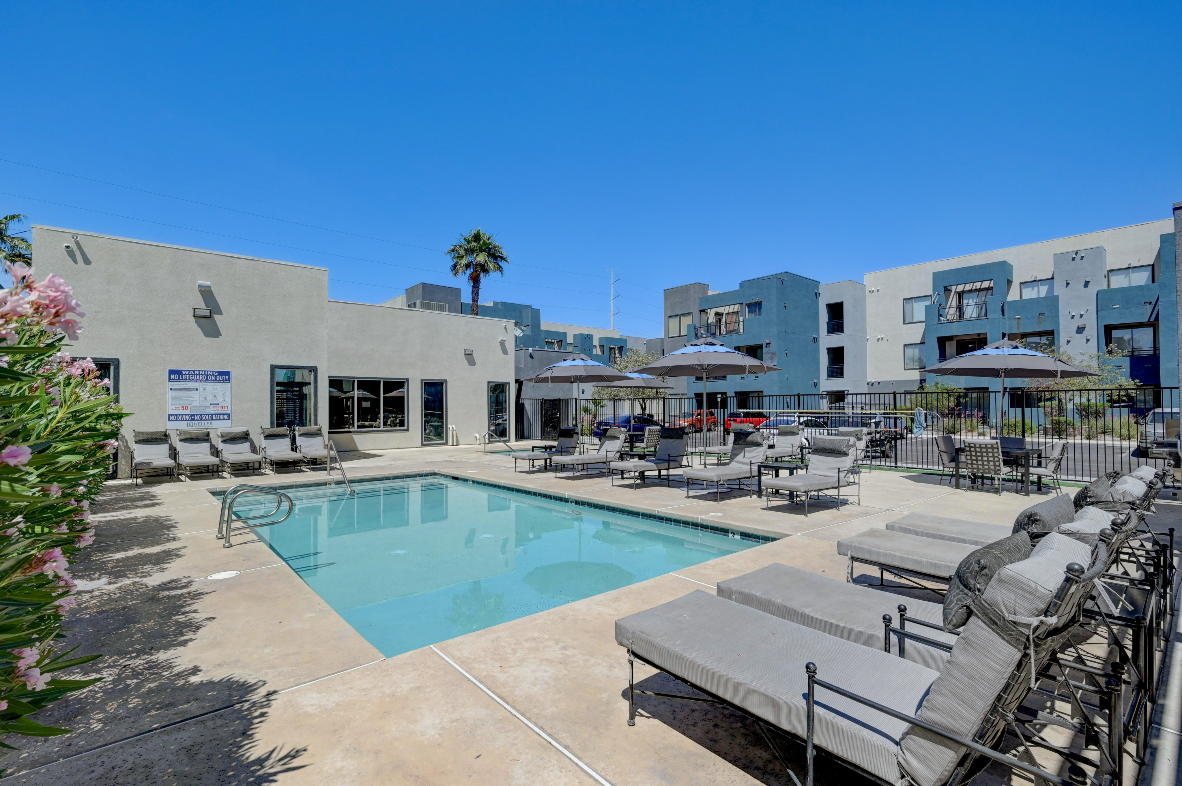 Inviting outdoor pool area with modern loungers and umbrellas, surrounded by contemporary apartment buildings under a clear blue sky.