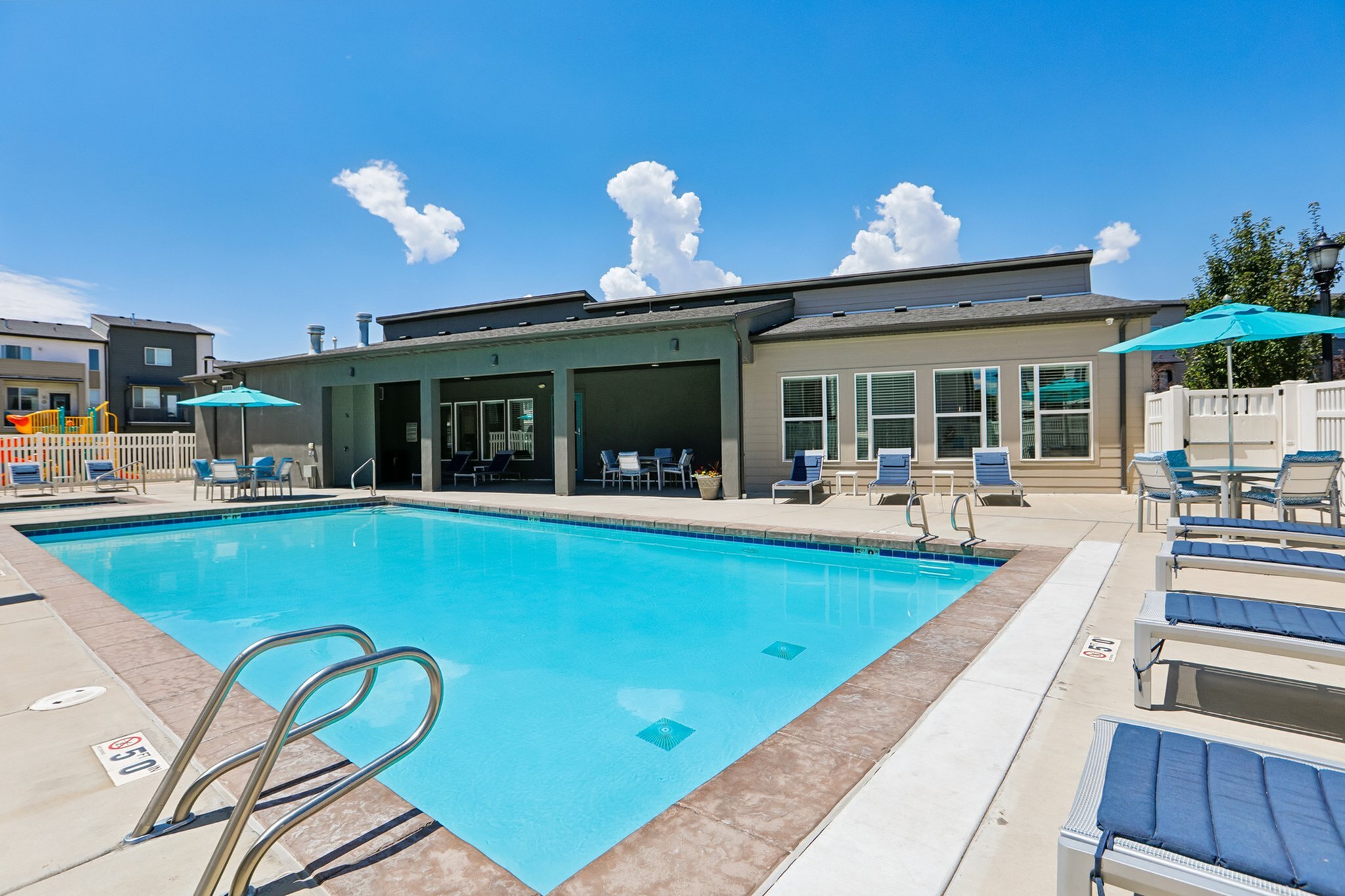 Inviting swimming pool area with sun loungers, shaded seating, and modern clubhouse under a clear blue sky.