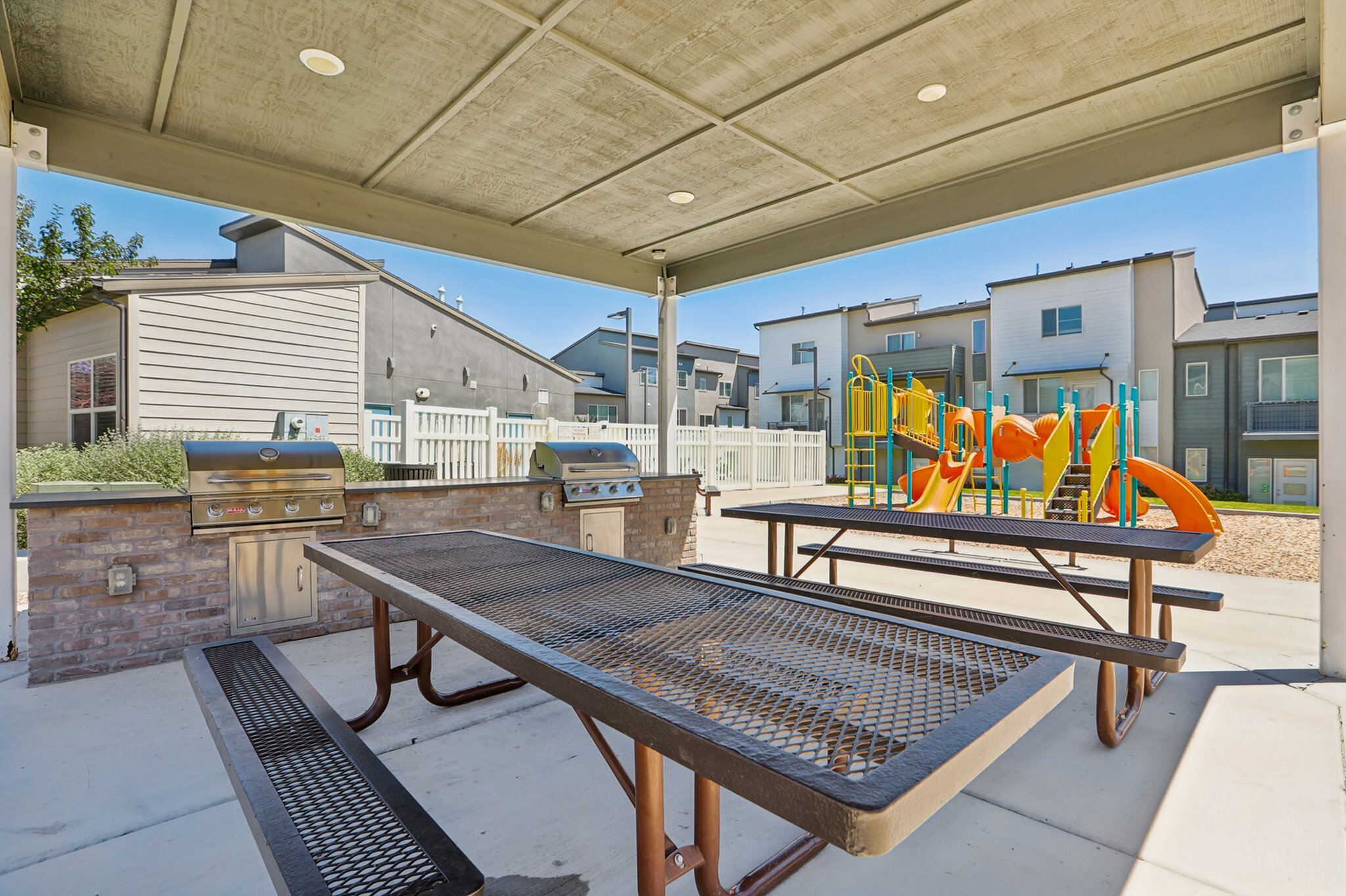 Charming outdoor patio area with grills and picnic tables beside a vibrant playground, perfect for family gatherings in this modern apartment community.
