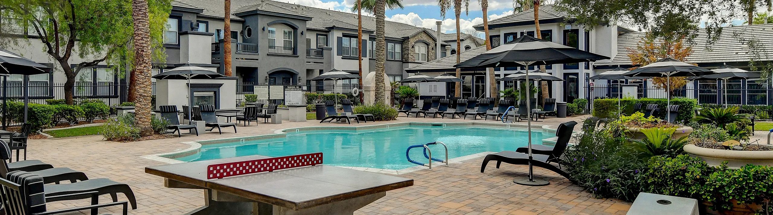 Luxurious apartment community featuring a sparkling swimming pool, surrounded by elegant lounge chairs and lush landscaping under a vibrant blue sky with palm trees.
