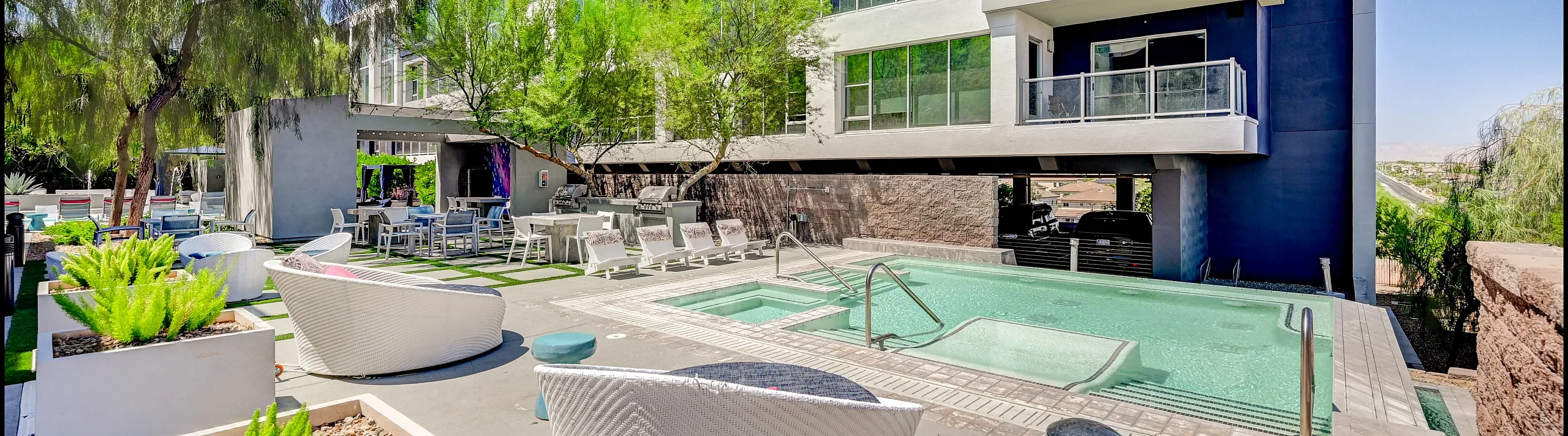 Modern apartment complex with luxurious pool area, stylish outdoor seating, and lush landscaping under a bright blue sky.